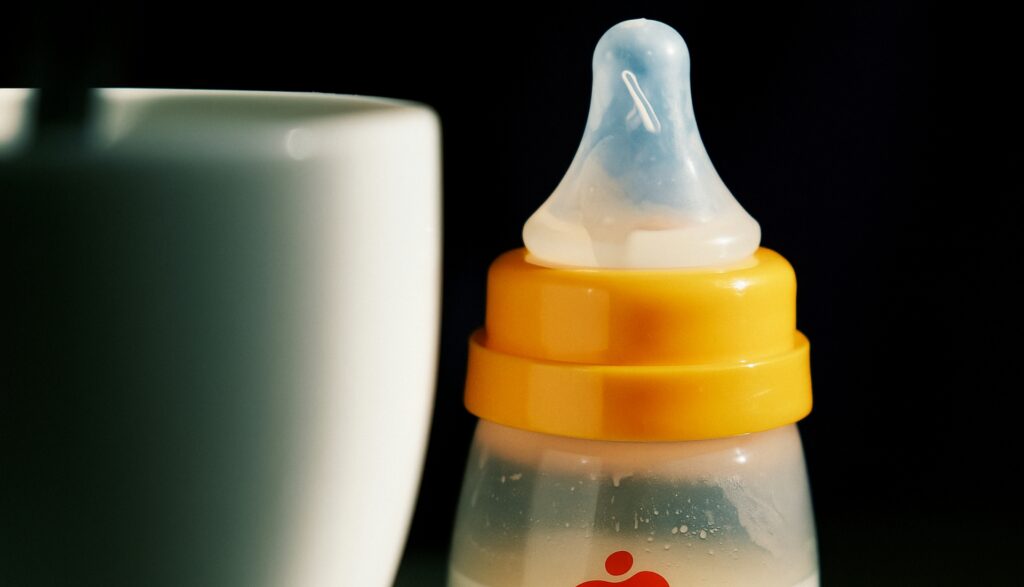 Primer plano de un biberón de plástico con tapa amarilla y tetina de silicona, parcialmente lleno de leche junto a una taza blanca desenfocada.
