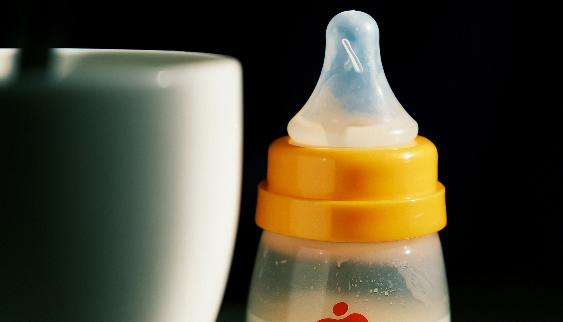 Primer plano de un biberón de plástico con tapa amarilla y tetina de silicona, parcialmente lleno de leche junto a una taza blanca desenfocada.