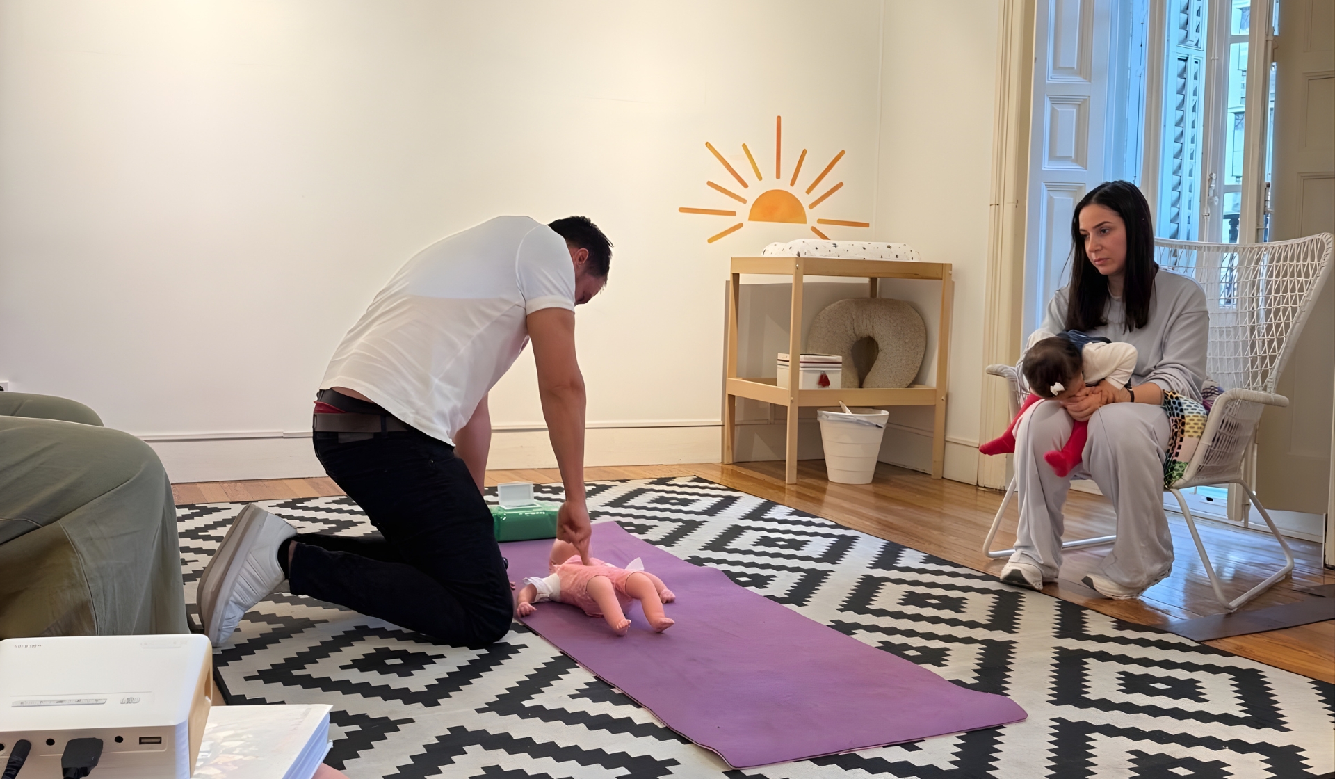 Hombre arrodillado practica maniobras de primeros auxilios sobre un muñeco de bebé en una esterilla mientras una madre con su hija en brazos observa sentada.