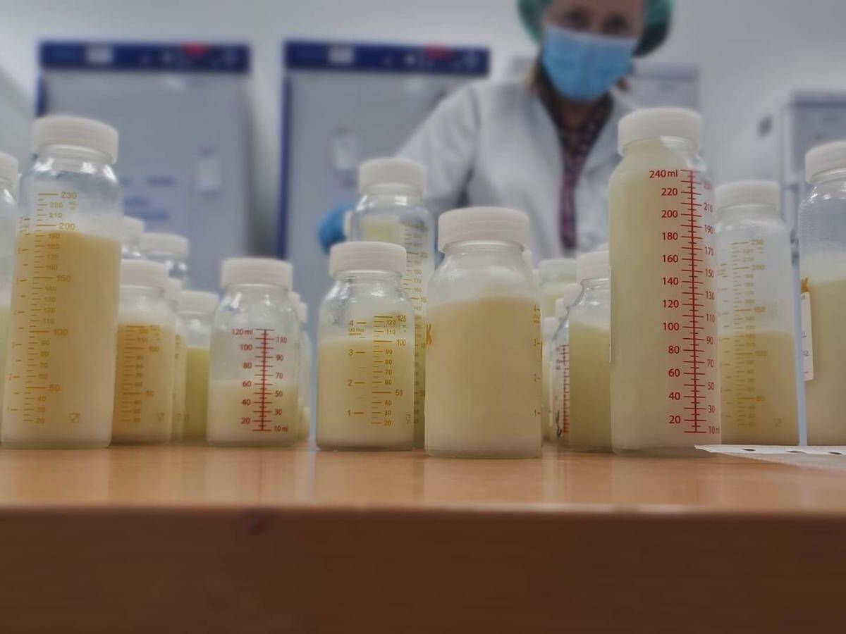 Varios biberones transparentes llenos de leche materna alineados sobre una mesa en una sala clínica, con una profesional sanitaria desenfocada al fondo.