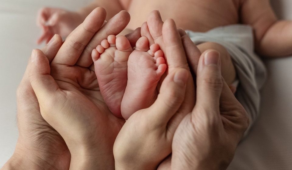 Manos de dos adultos sostienen los pies descalzos de un bebé recién nacido tumbado.