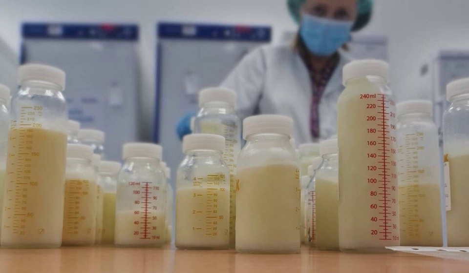 Varios biberones transparentes llenos de leche materna etiquetados y alineados sobre una mesa en una sala de laboratorio, con una profesional sanitaria desenfocada al fondo.