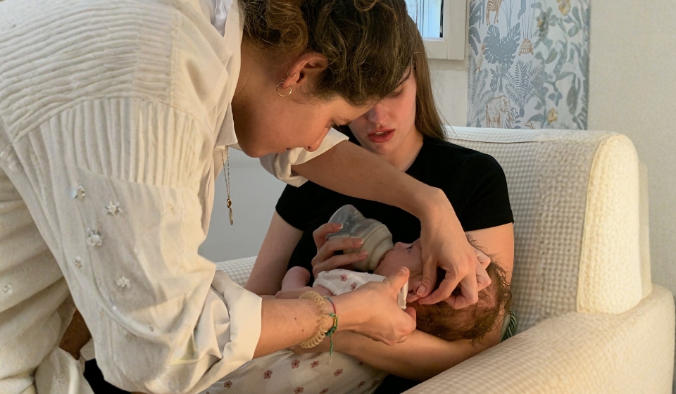 La matrona Elena Tovar Muñoz coloca los primeros pendientes a un bebé mientras la madre lo sostiene y le da el biberón en un sillón.