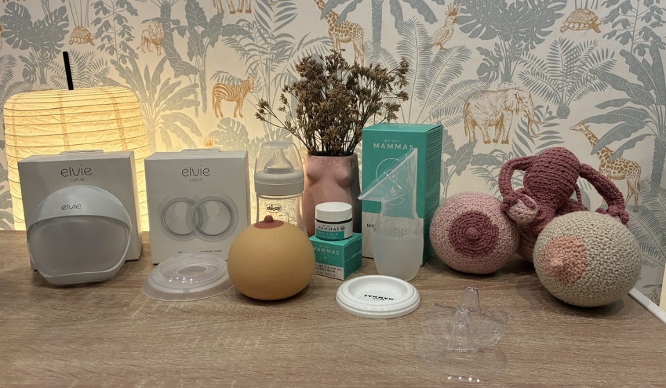 Mesa de madera con sacaleches, biberón, pechos de ganchillo y productos de cuidado del pecho iluminados por una lámpara cálida.