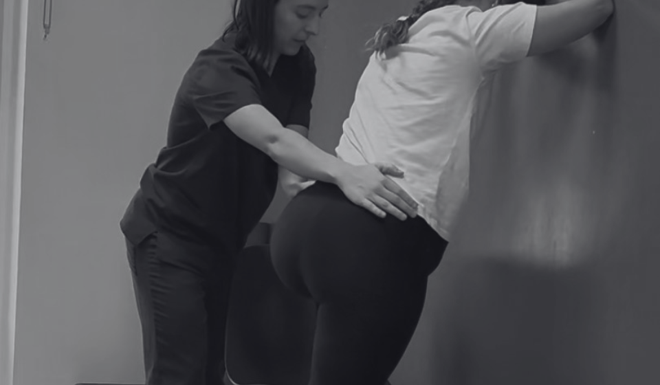 Matrona y fisioterapeuta de suelo pélvico realizando ejercicios asimétricos para el pródromos de parto contra pared.